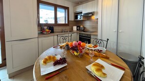 Uma cozinha moderna com armários brancos, uma mesa de jantar com croissants e frutas, e vista para o exterior.