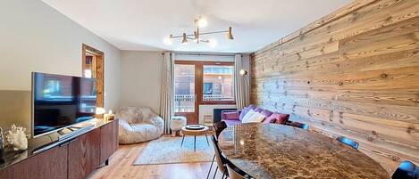 A modern living room with a large flat-screen TV, a wooden dining table with chairs, a sofa, and a wooden accent wall.