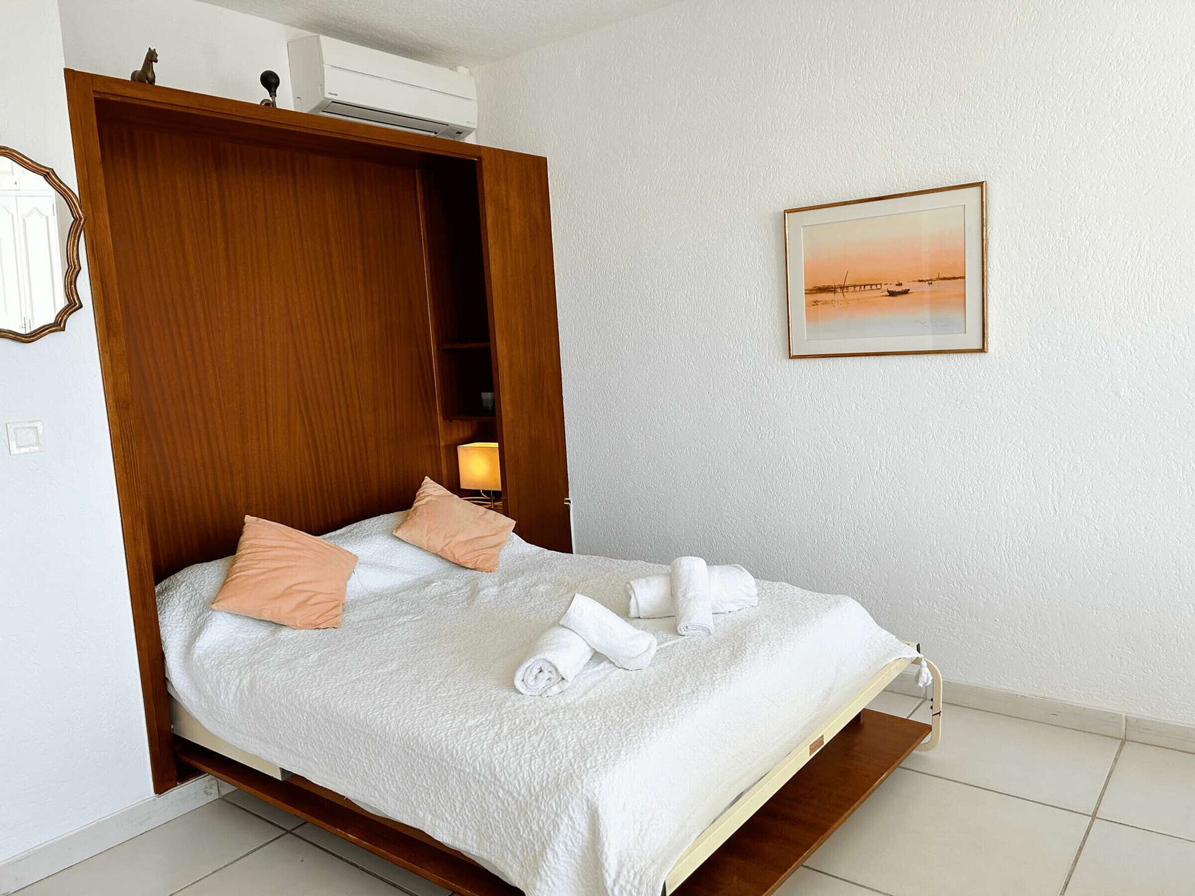 A bedroom with a bed, a wooden wardrobe, a nightstand with a lamp, and a framed picture on the wall.