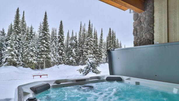 A hot tub with a stone wall backdrop, surrounded by snow-covered trees. - Bottom Floor, Walk Out Condo with great ski access and private hot tub! (Sun Peaks)
