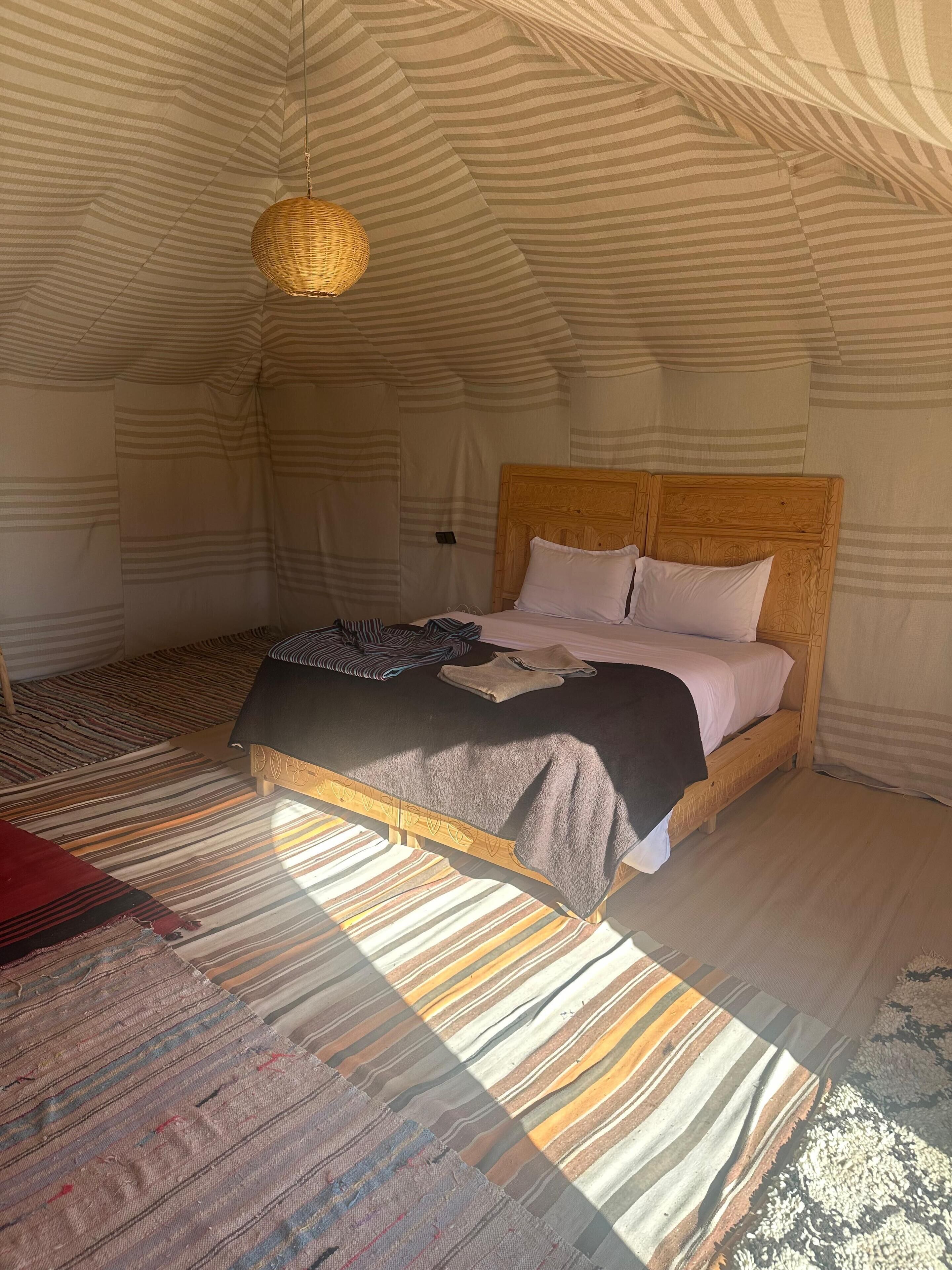 A cozy, striped-patterned tent with a wooden bed, a woven light fixture, and a patterned rug.