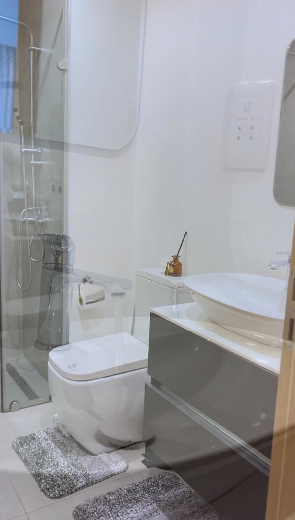 A modern bathroom with a glass shower enclosure, a white toilet, a sink with a mirror, and a small shelf with a bottle. - Experience the height of Accra luxury in the prestigiousNova at Roman Ridge. (Accra)