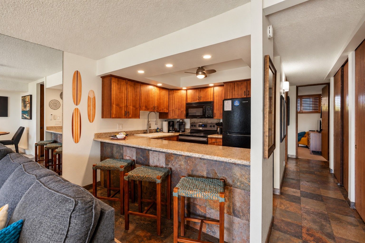 A modern kitchen with wooden cabinets, a black refrigerator, and a granite countertop. There are three bar stools with woven seats.