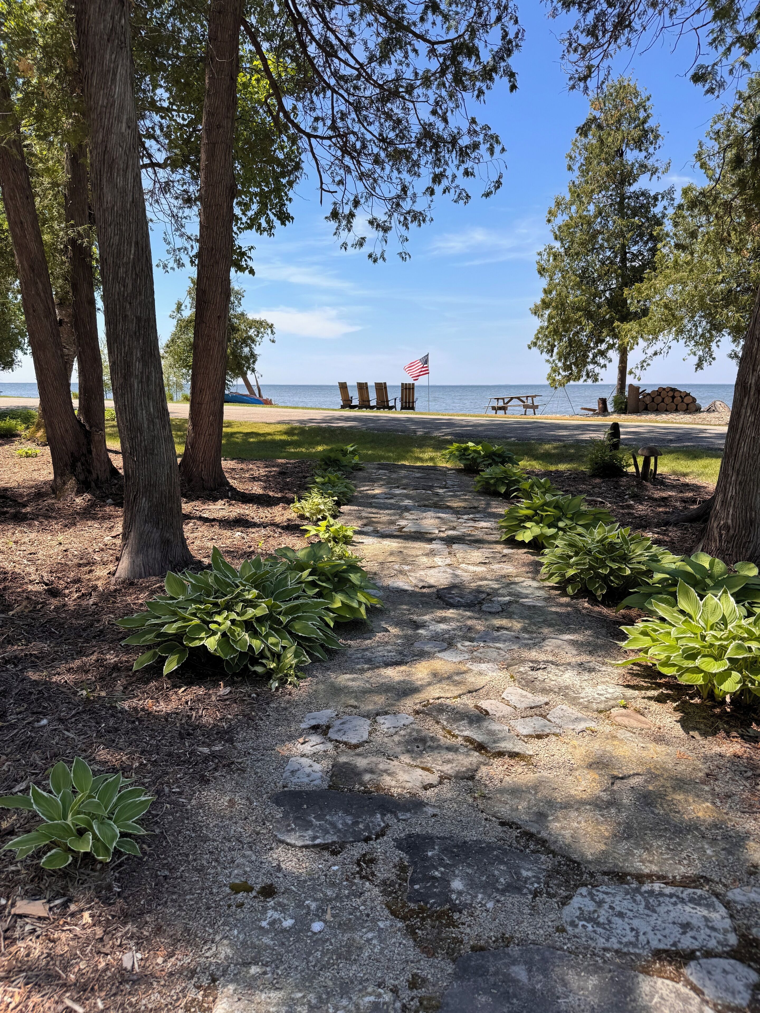Un chemin de pierres menant à des chaises de plage et à un drapeau, le tout entouré d’arbres et de plantes.