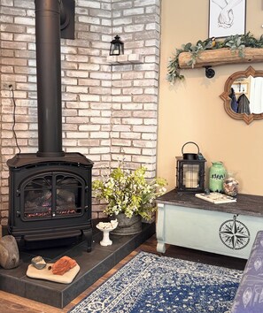 A cozy living room with a brick wall, a black wood-burning stove, a small table with a bowl of snacks, a potted plant, and a blue patterned rug.