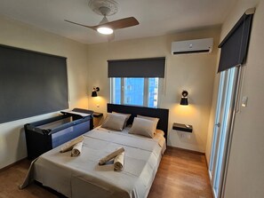 Ein Hotelzimmer mit Bett, Deckenventilator, Klimaanlage und Fenster mit Blick auf Gebäude.