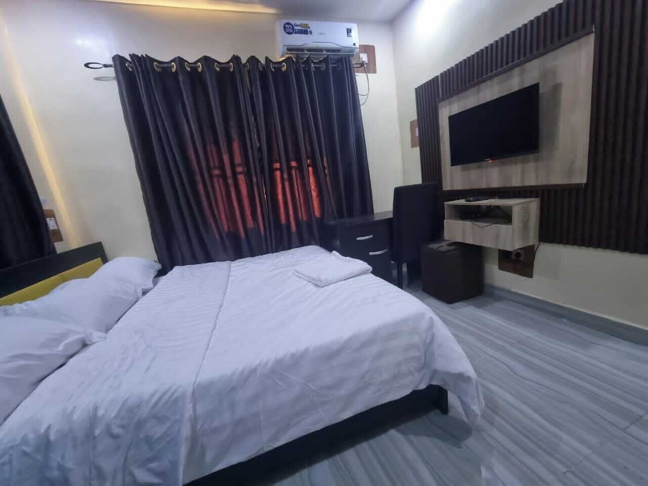 A hotel room with a bed, a desk, a chair, a TV, and an air conditioning unit.