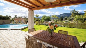 An outdoor patio with a wooden table and chairs, a stone pillar, and a view of a swimming pool and mountains in the background. - Villa del Borgo-Private pool, exclusive park, authenticity and luxury by the sea (Sardegna)