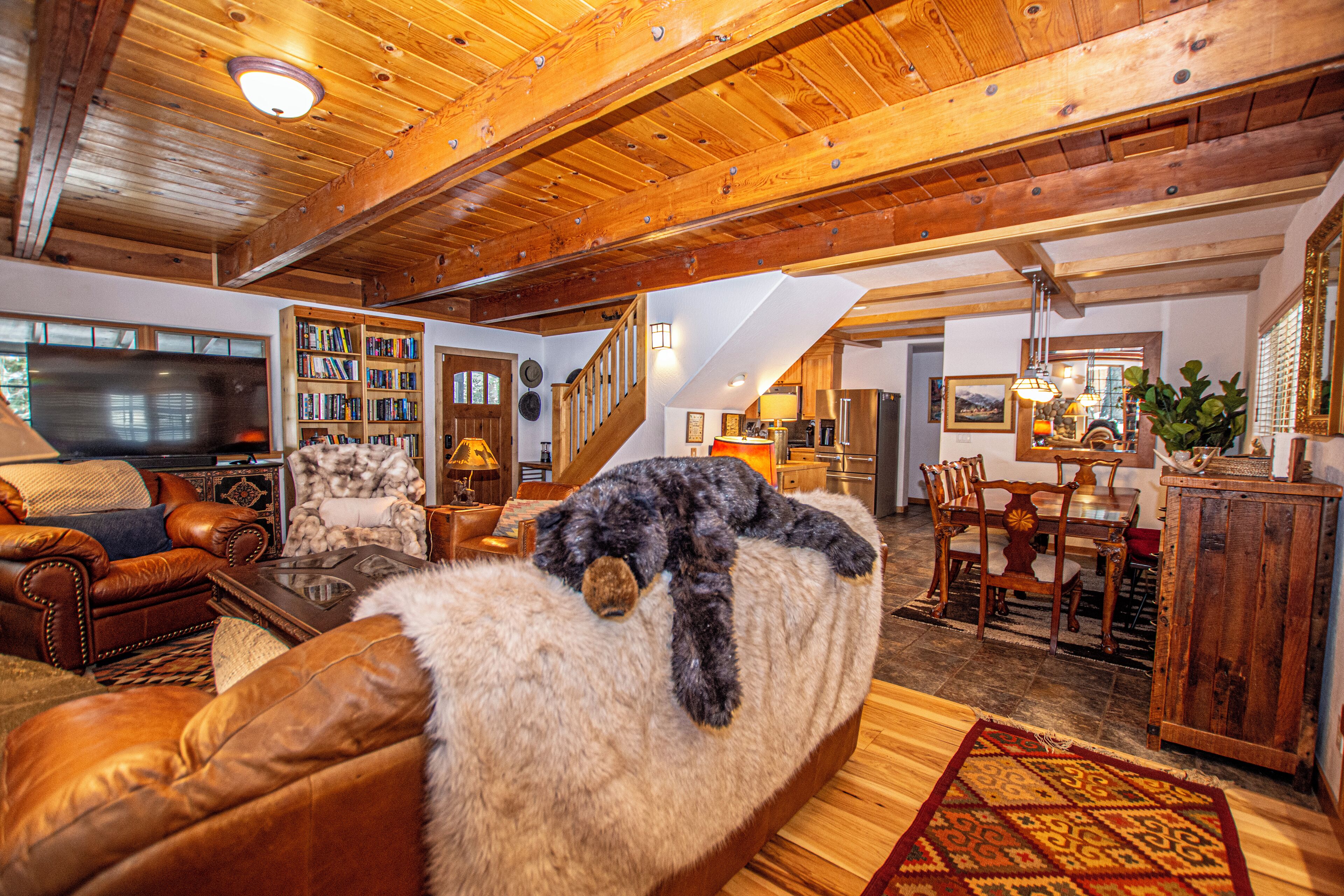 Ein gemütliches Wohnzimmer mit einem Hund auf einem fellbezogenen Hocker, Holzbalken an der Decke, einer Treppe und einem Essbereich.