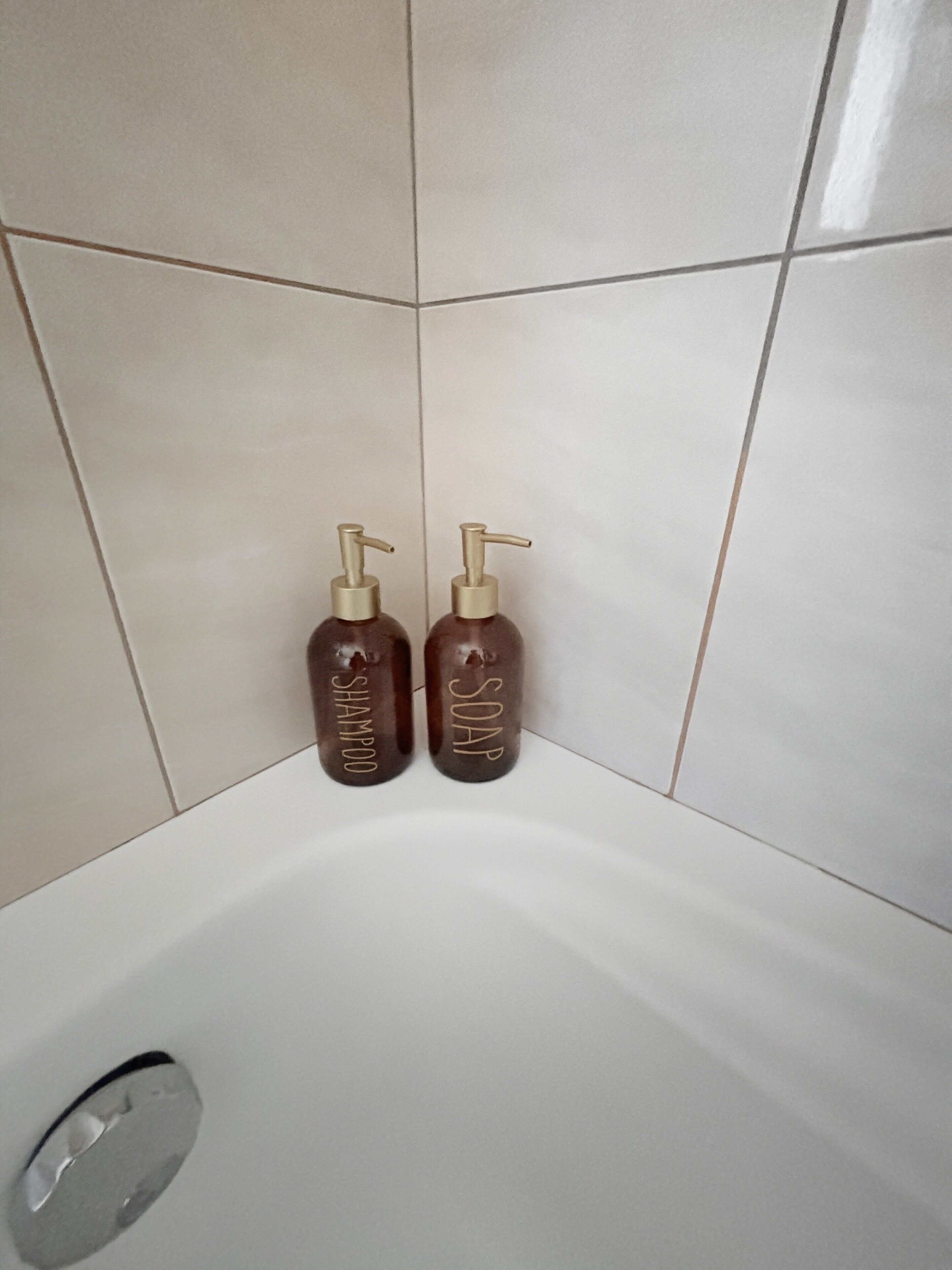 Baño con dos botellas marrones etiquetadas 'SHAMPOO' y 'SOAP' sobre una pared de azulejos blancos.