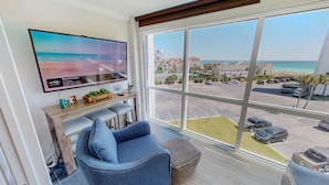 A living room with a large window, a flat-screen TV, a sofa, and a small wooden table with plants. - Shoreline Towers 3054 2 Bedroom Condo (Destin)