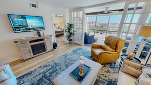 A living room with a television, sofa, armchair, and a view through large windows. - Shoreline Towers 3054 2 Bedroom Condo (Destin)