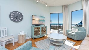 A living room with a large window overlooking a beach, a round coffee table, a sofa, and a television. - Phoenix V 115 2 Bedroom Condo (Orange Beach)