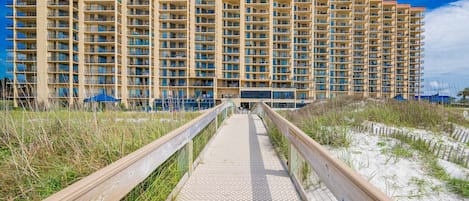 A high-rise hotel with multiple balconies, a wooden boardwalk leading to the entrance, and a sandy beach area.