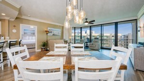 A spacious dining area with a wooden table and white chairs, a ceiling fan, and large windows offering a city view. - Phoenix On The Bay 1511 3 Bedroom Condo (Orange Beach)