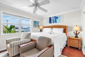 Une chambre d’hôtel avec un lit, deux chaises, une table de chevet, une lampe, un ventilateur de plafond et une grande fenêtre donnant sur un beau paysage.