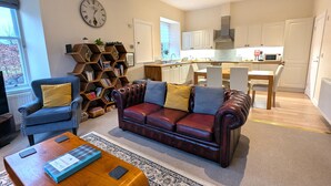 Un salon avec un canapé en cuir, des fauteuils, une table basse et une bibliothèque. On aperçoit une cuisine au fond.