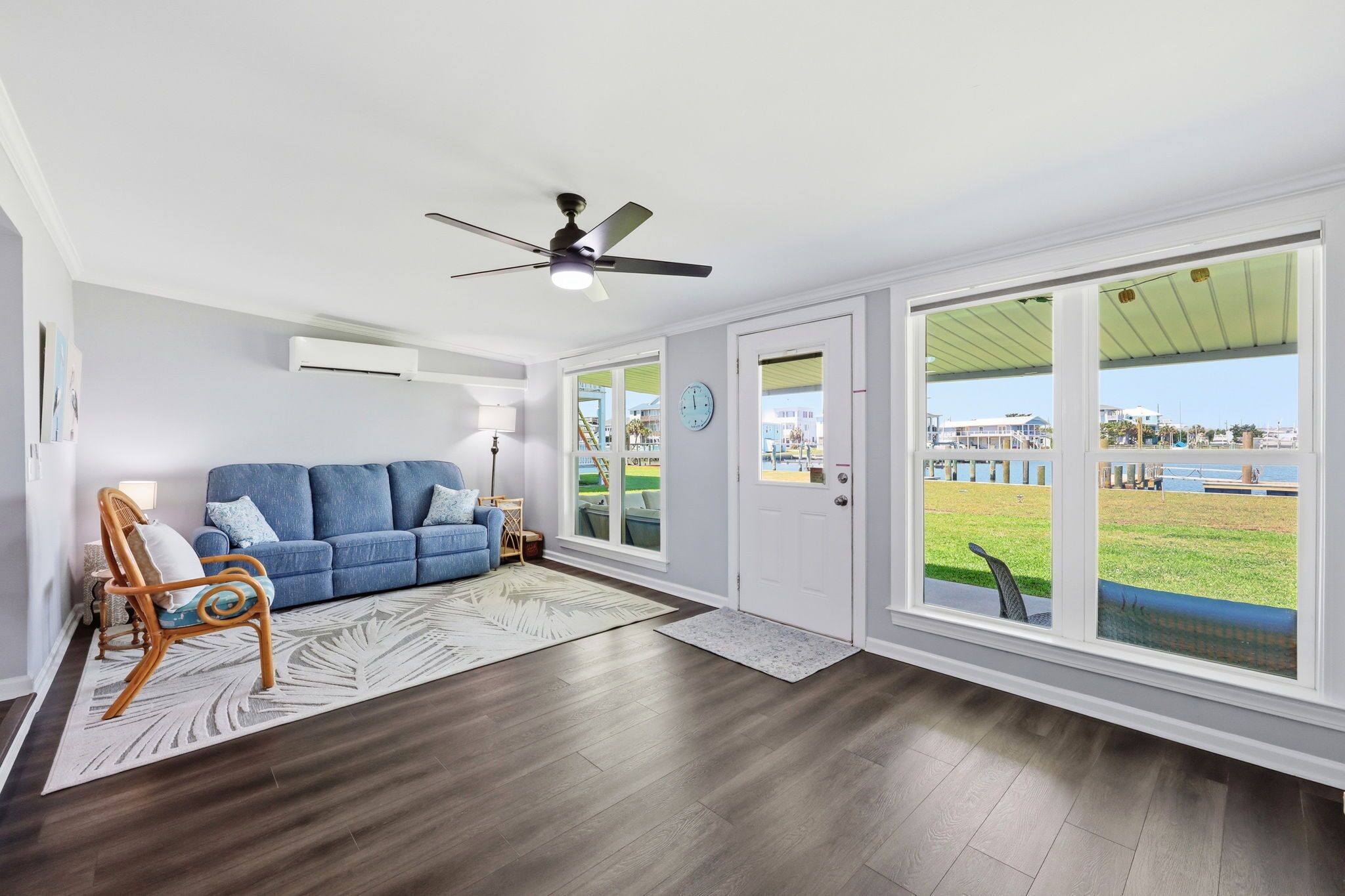 A spacious living room with a blue sofa, a wooden chair, a ceiling fan, and large windows with a view of a grassy area and buildings.
