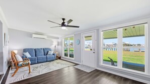Ein geräumiges Wohnzimmer mit einem blauen Sofa, einem Holzstuhl, einem Deckenventilator und großen Fenstern mit Blick auf eine Rasenfläche und Gebäude.
