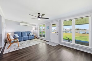 A spacious living room with a blue sofa, a wooden chair, a ceiling fan, and large windows with a view of a grassy area and buildings. - Bayview Anchor-Pet Friendly Home with Dock (Atlantic Beach)