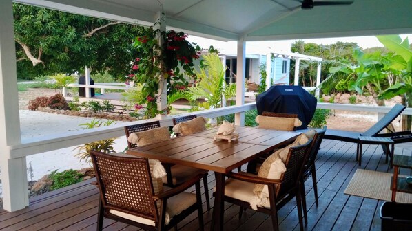 A covered outdoor dining area with a wooden table and wicker chairs, surrounded by lush greenery and a view of a house.