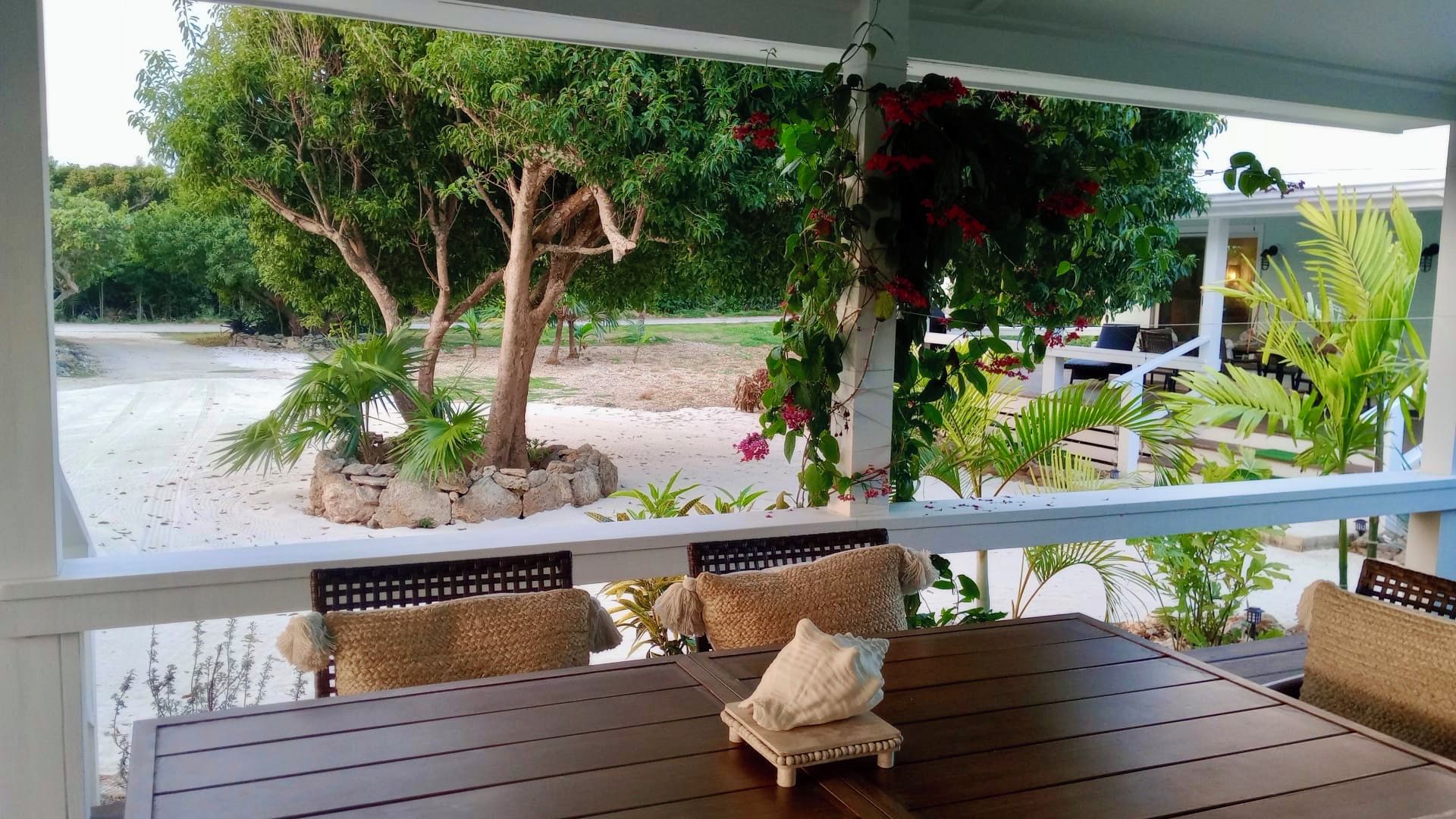 Un espace extérieur abrité, aménagé avec une table et des chaises en bois, entouré d’une végétation luxuriante et offrant une vue sur un paysage sablonneux.