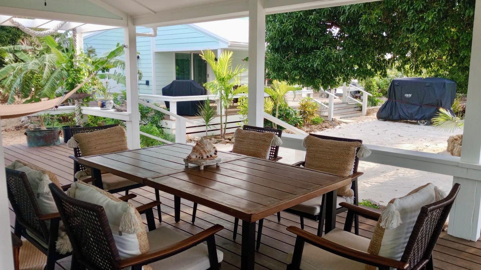 A covered outdoor dining area with a wooden table and wicker chairs, surrounded by lush greenery and a view of a blue house in the background.