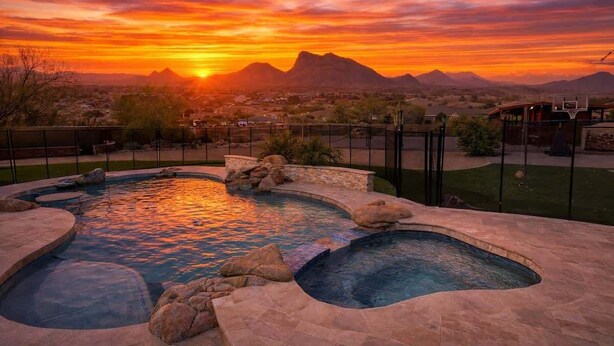 A swimming pool with a curved design, surrounded by stonework and rocks, set against a sunset with mountains in the background. - The Family Room-Pool & Outdoor Games-Game Room (Scottsdale)