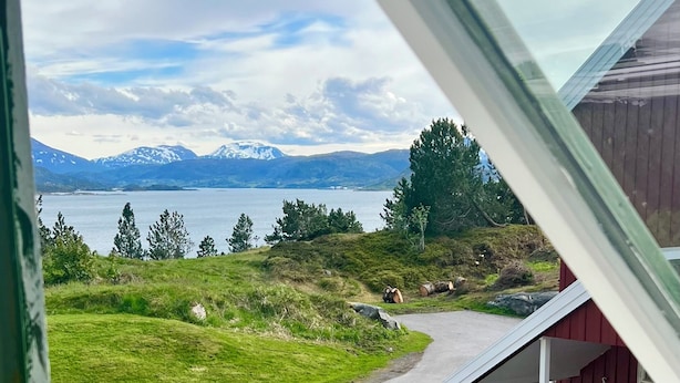 Vista a través de una ventana de un paisaje montañoso con un lago, árboles y un edificio rojo.