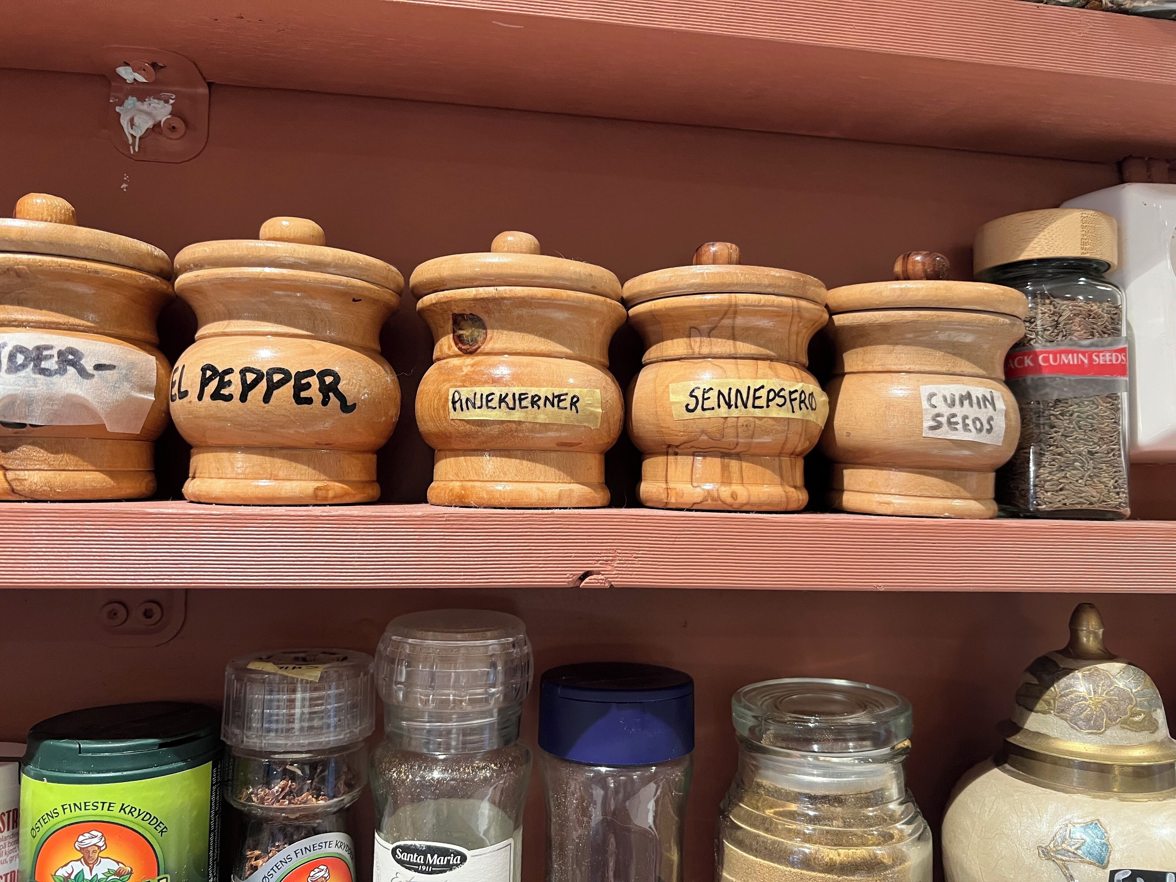Une étagère avec des pots à épices en bois, chacun portant une étiquette indiquant son nom, comme « EL PEPPER » et « ANJEKJIERNER ».