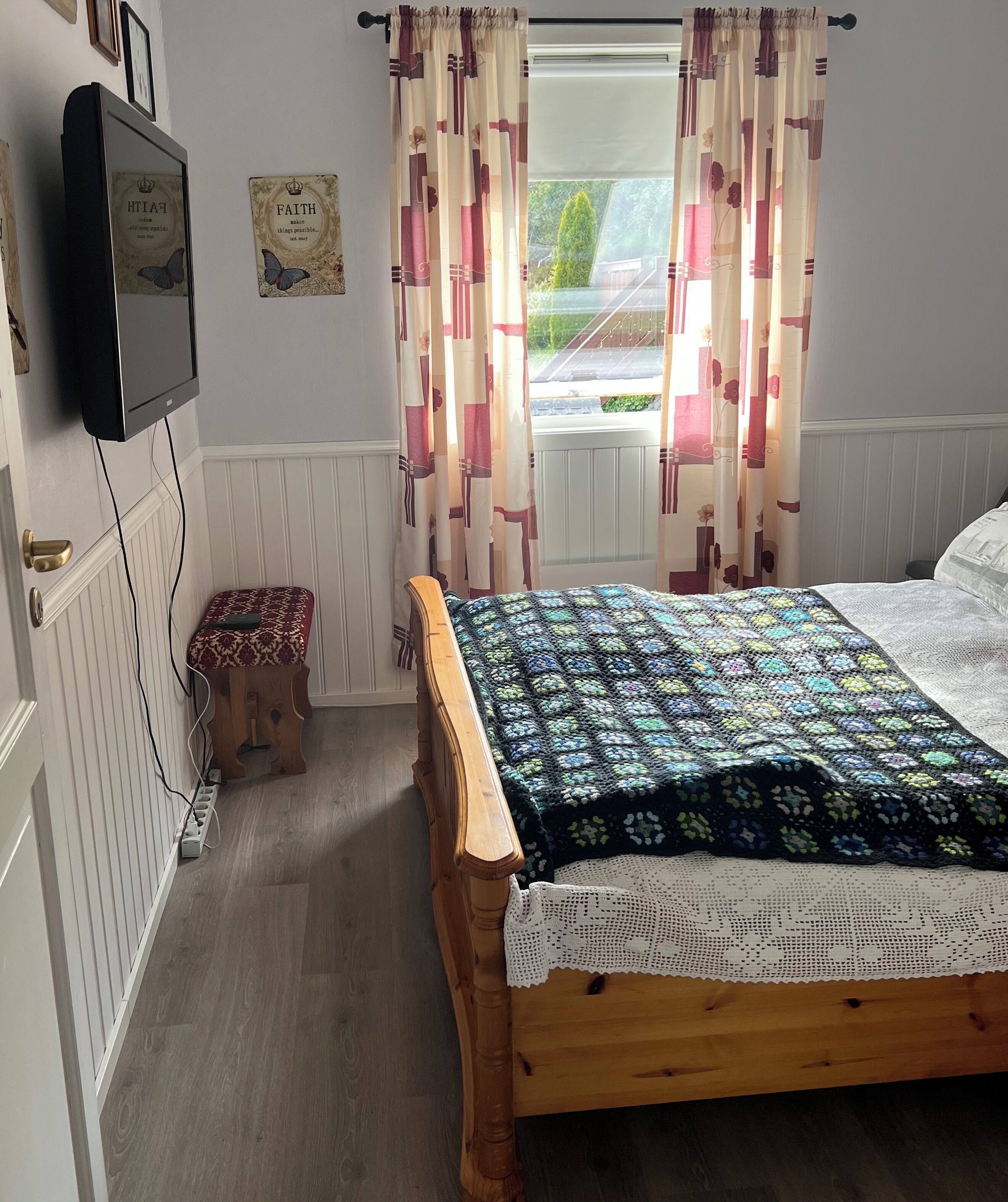 A bedroom with a wooden bed, a quilt, a small stool, a television, and a window with curtains.