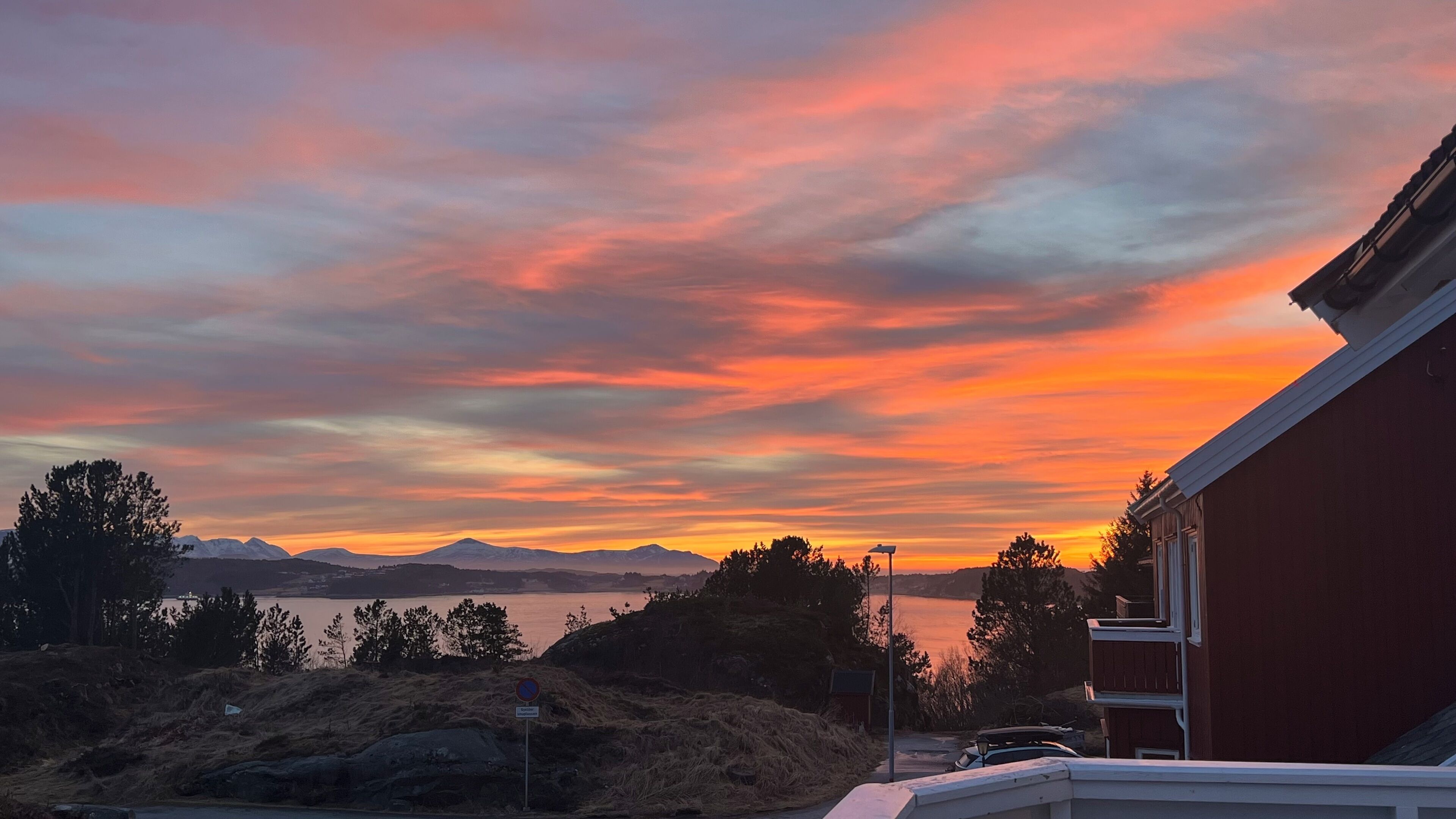 A sunset over a mountainous landscape with a body of water, a building with a red exterior, and a no parking sign.