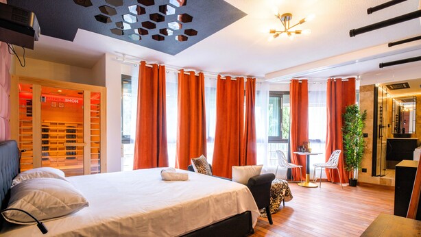 Une chambre d’hôtel moderne avec un grand lit, des rideaux orange, un coin salon avec une table et des chaises, et un magasin visible appelé « MOMENTX ».