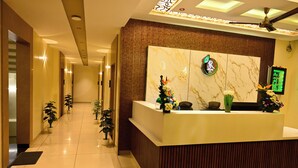 A reception area with a marble reception desk, a peacock logo, and a mirror with lights. There are potted plants along the corridor.