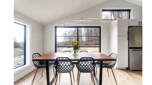 Un comedor moderno con una mesa de madera y sillas negras, un florero con flores amarillas y vistas a árboles a través de amplios ventanales.