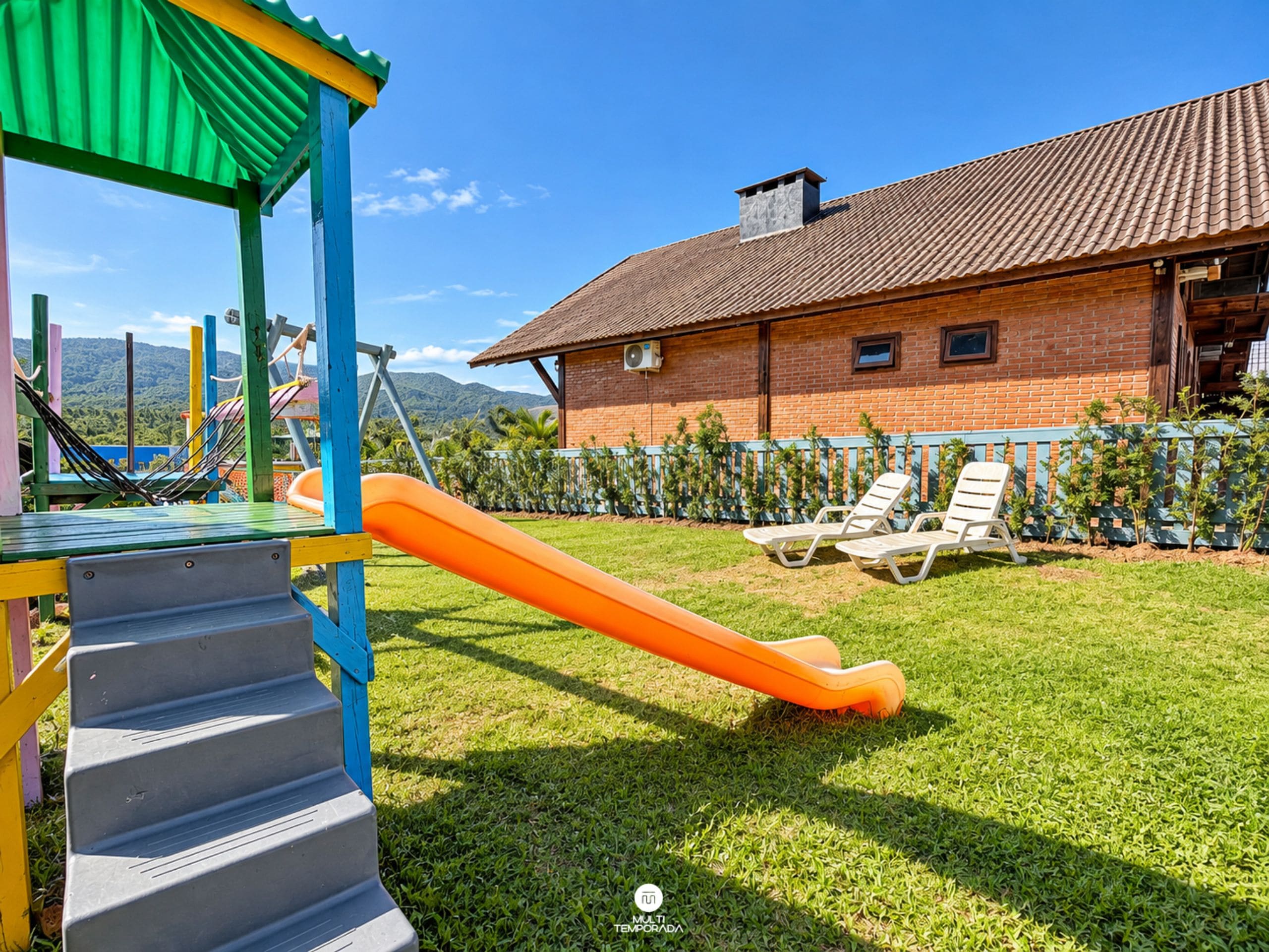 A playground with a slide and steps, a brick house with a tiled roof, and a fenced area with lounge chairs.