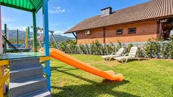 Un parque infantil con un tobogán y escalones, una casa de ladrillo con tejado de tejas, y un área cercada con sillas de descanso.