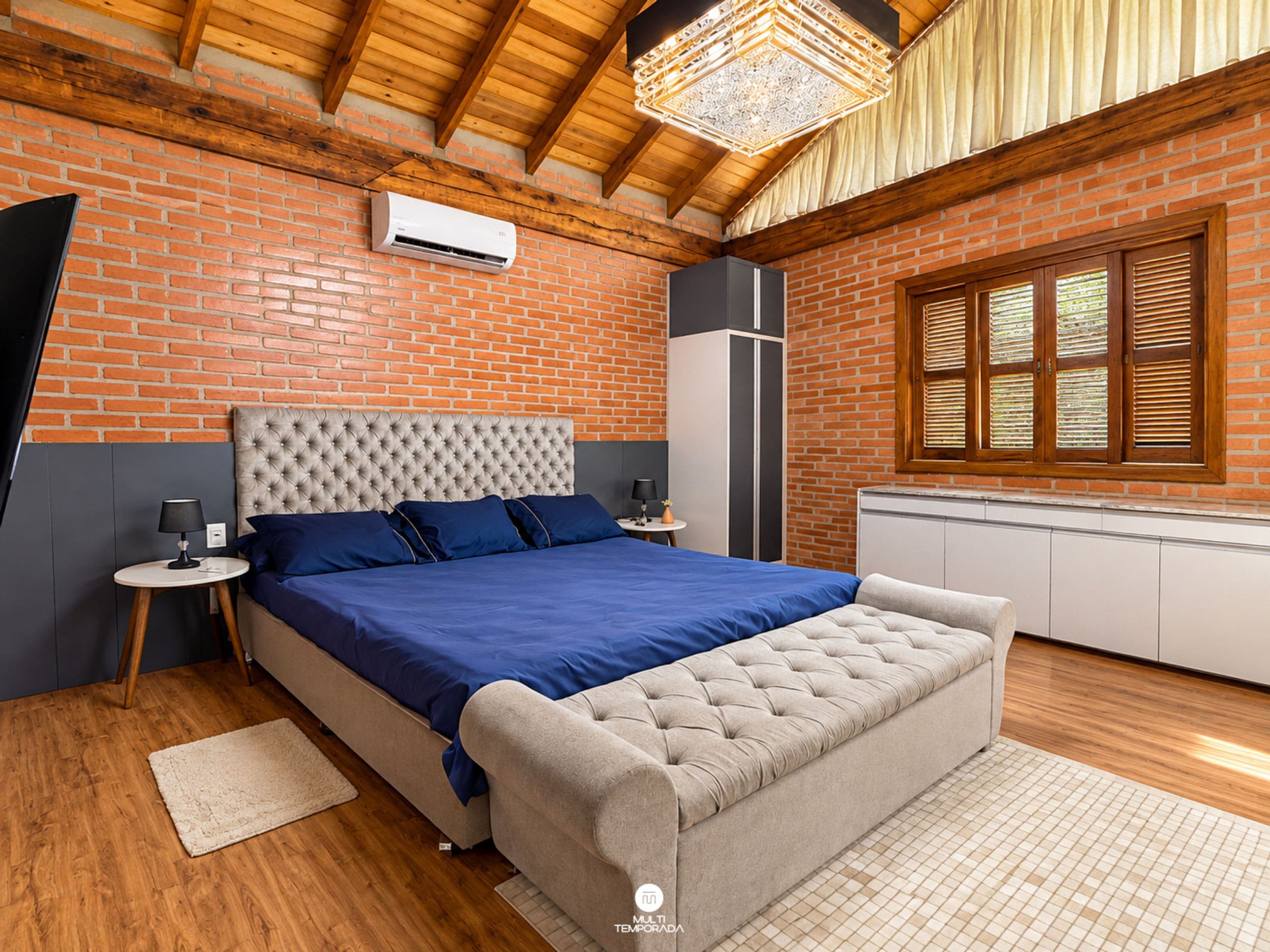 A bedroom with a brick wall, a wooden ceiling, a bed with blue bedding, a grey upholstered bench, and a window with wooden shutters.