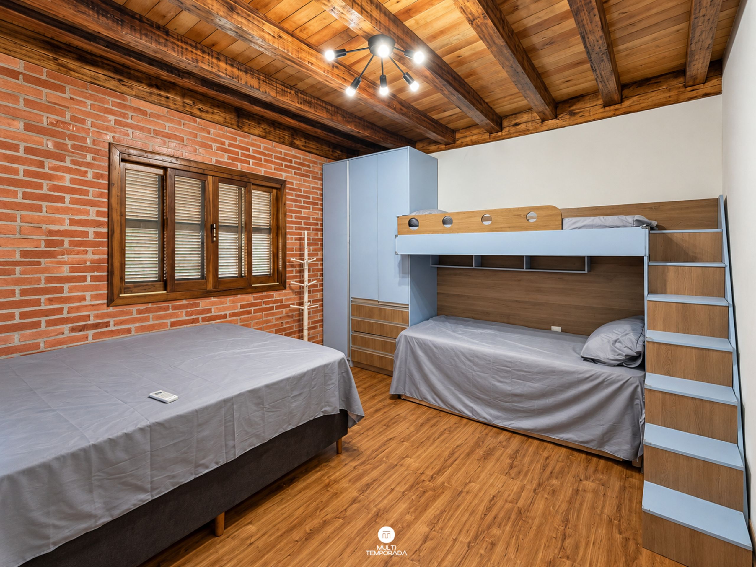 Una habitación con pared de ladrillo, vigas de madera en el techo, una cama litera y una cama individual.