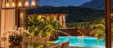A modern outdoor dining area with a glass table and chairs, overlooking a swimming pool and a mountainous landscape.