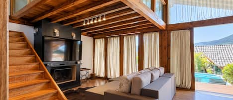 Un interior moderno con una escalera de madera, un amplio sofá modular, un televisor de pantalla plana y vistas a una piscina y montañas.
