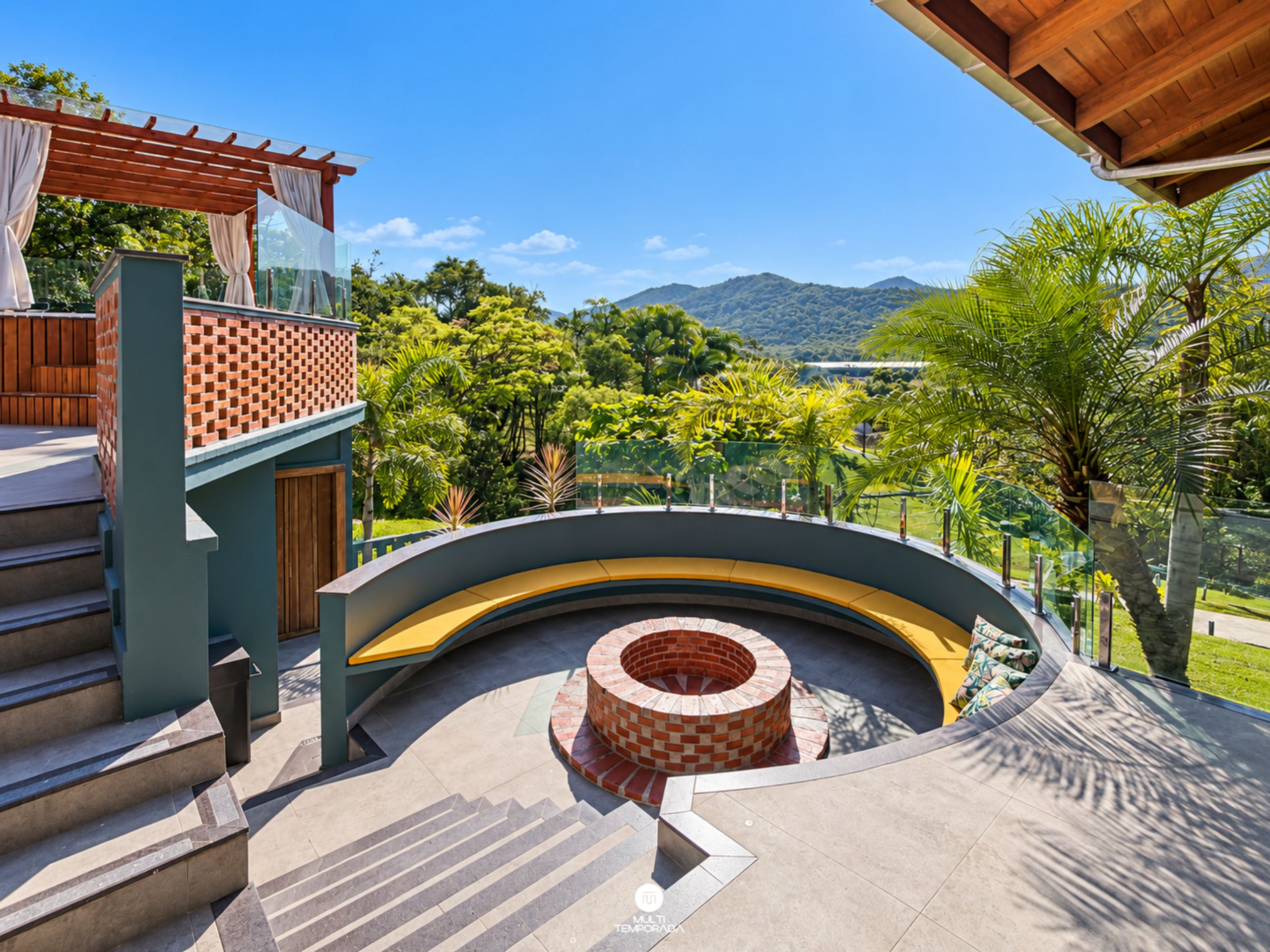 Un área exterior curva con una estructura de ladrillo, escalones que suben a una terraza de madera, y vistas a zonas verdes y colinas al fondo.