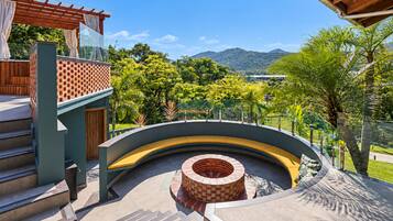Un área exterior curva con una estructura de ladrillo, escalones que suben a una terraza de madera, y vistas a zonas verdes y colinas al fondo.