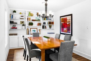 Un coin repas avec une table en bois, des chaises à revêtement gris, des meubles blancs et une bibliothèque remplie de livres et d’objets décoratifs.