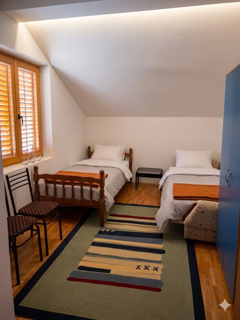 A small, cozy bedroom with two single beds, a wooden chair, a small table, and a window with blinds.