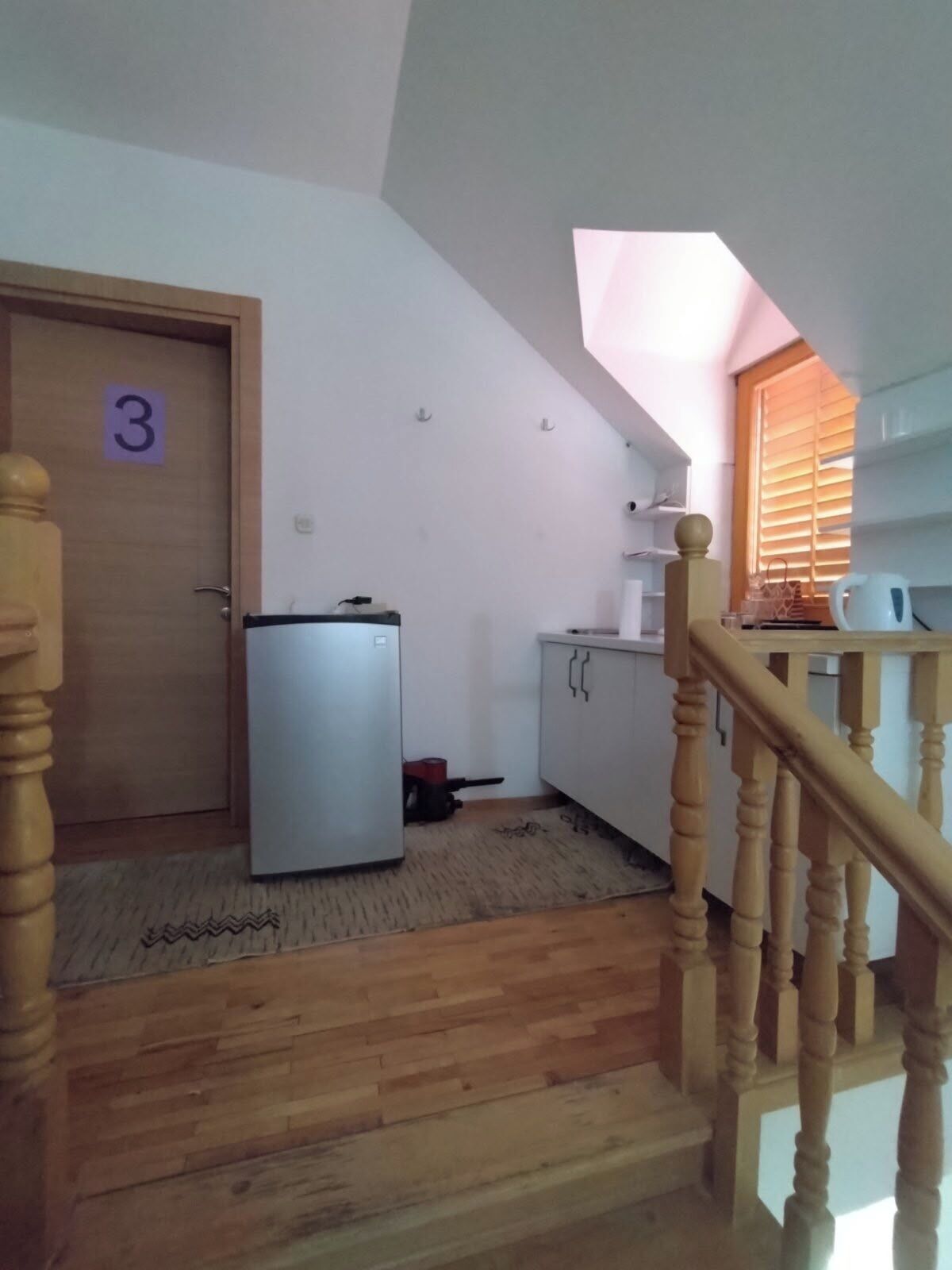 A room with a wooden staircase, a door marked with the number 3, a white refrigerator, a small heater, and a window with blinds.
