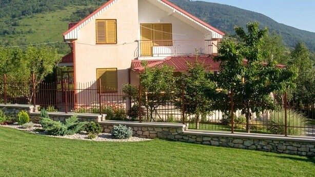 A two-story house with a red roof, a balcony, and a stone wall in front.