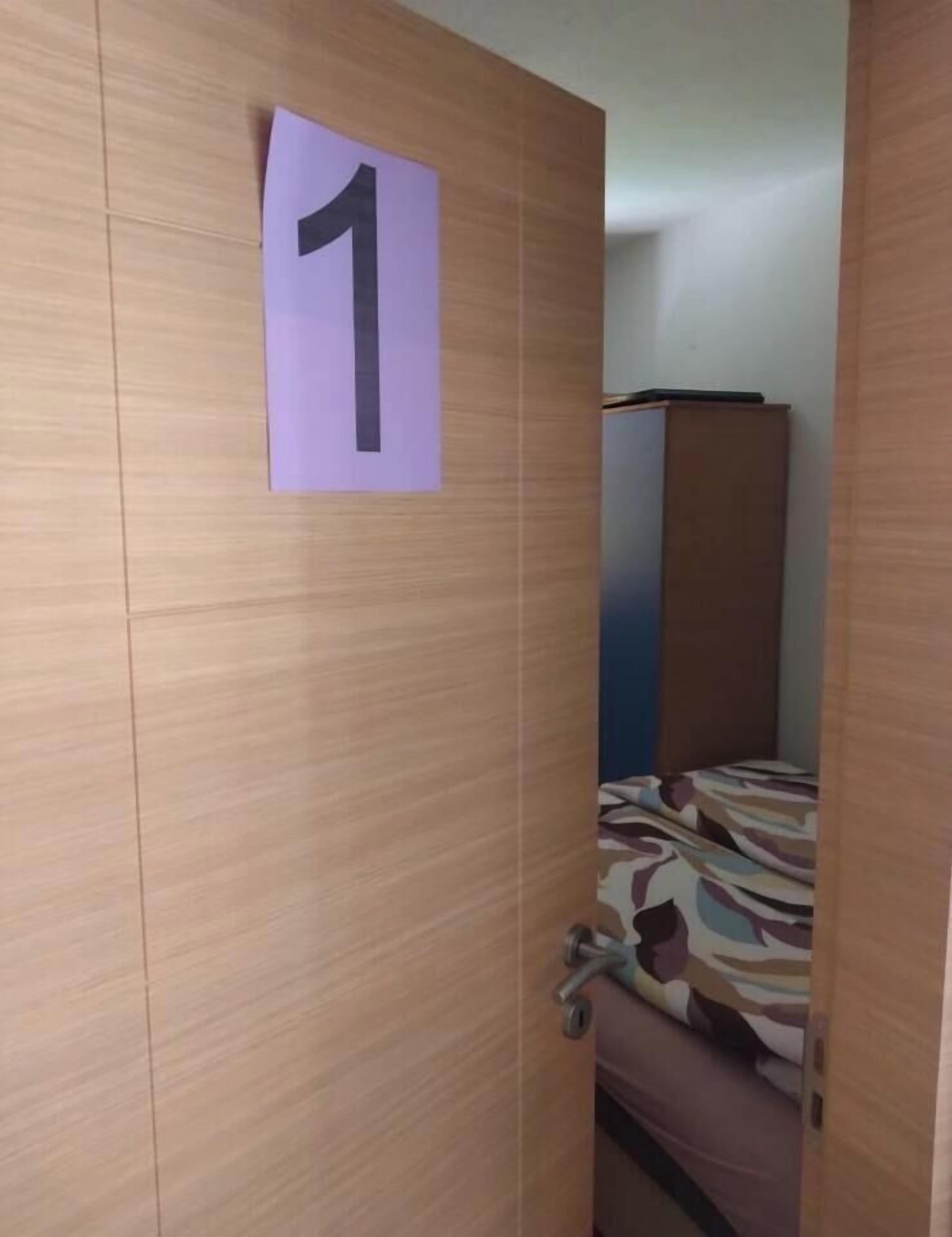 A room with a wooden door marked with the number 1, a bed with a patterned bedspread, and a small cabinet in the background.