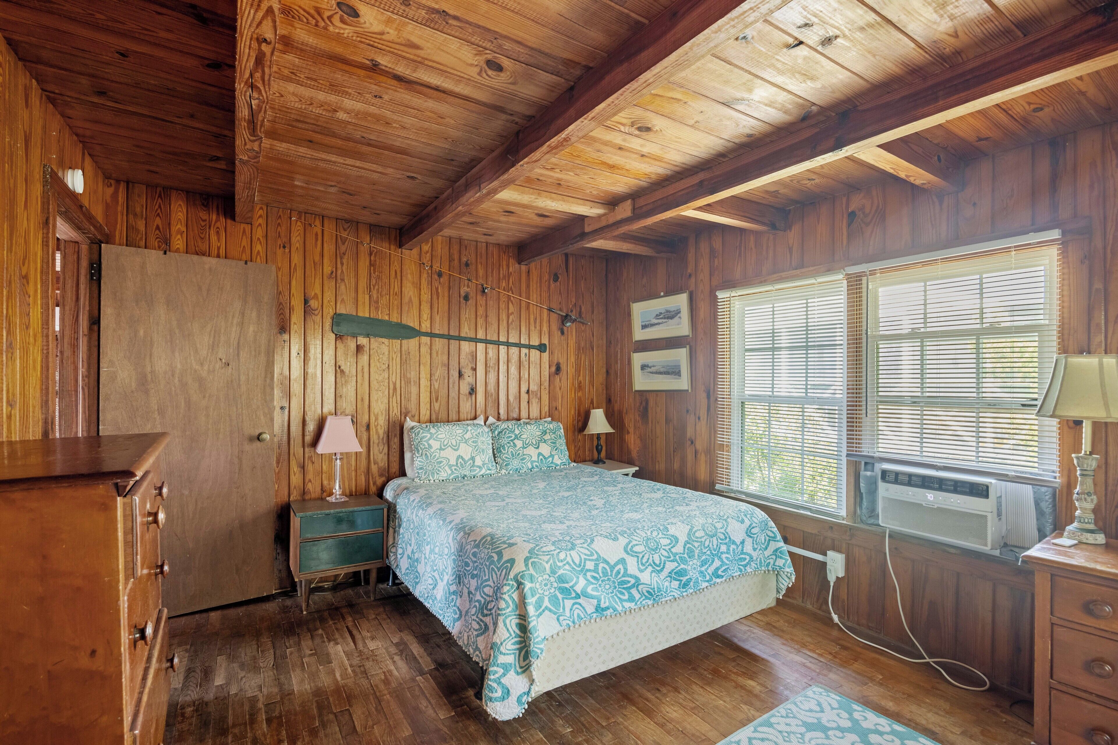 A wooden cabin bedroom with a bed, dresser, and a window with blinds.