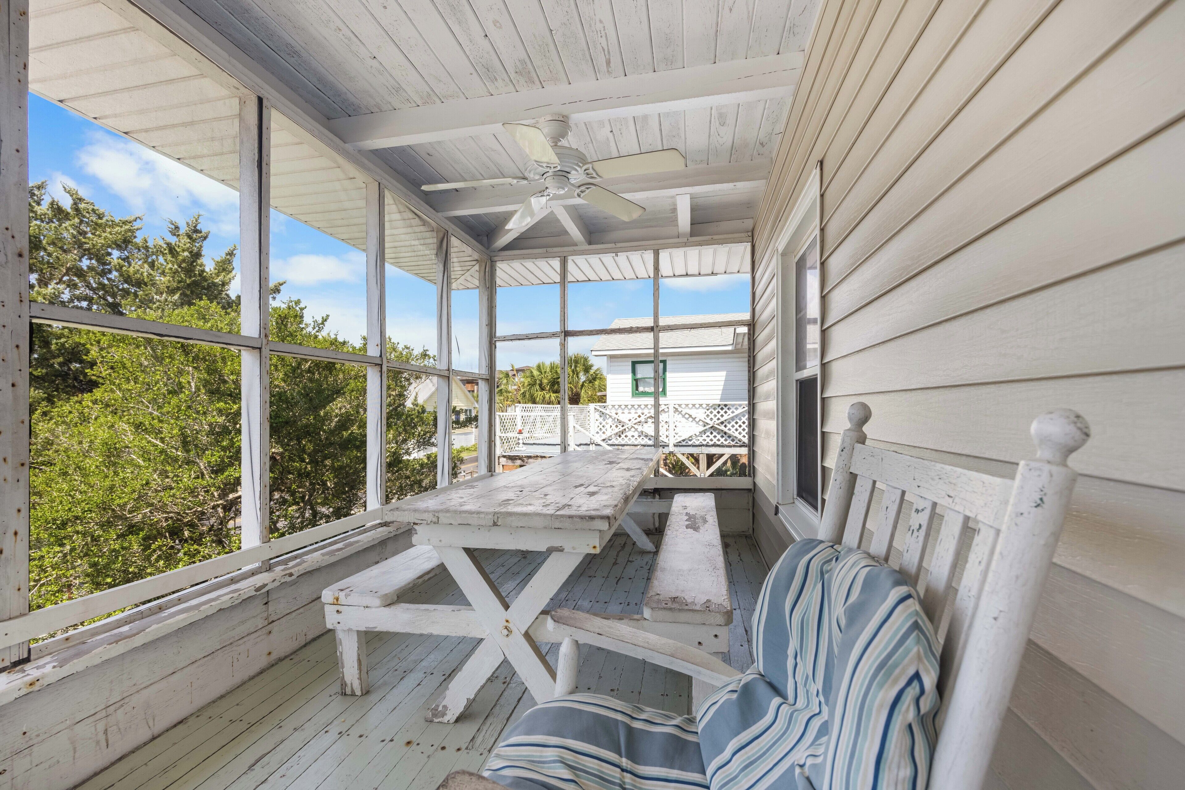 A covered porch with a wooden table and benches, large windows, and a ceiling fan.
